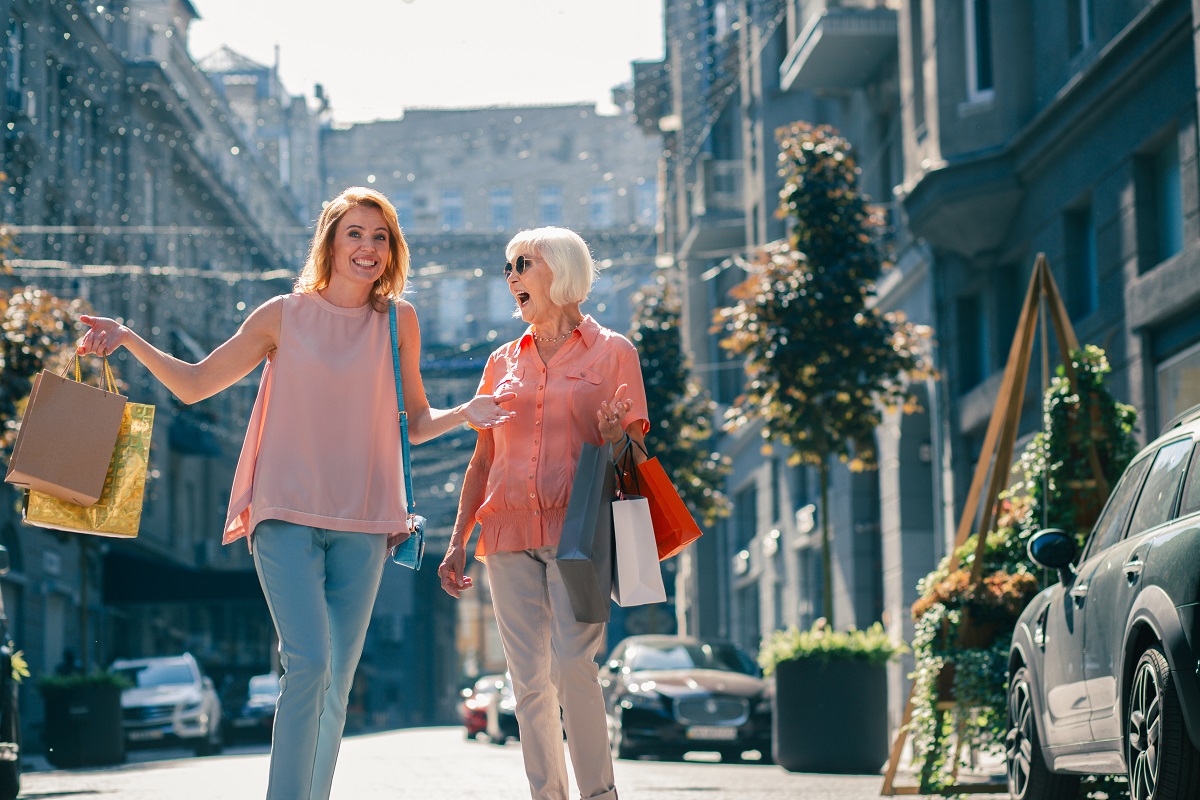 Positive adult lady and her retired mother enjoying shopping together and laughing with paper bags in hands