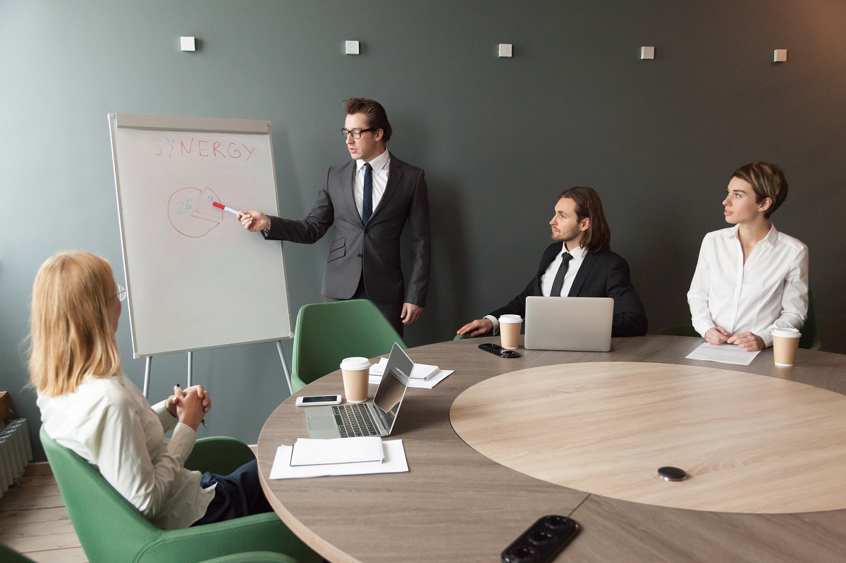 Confident speaker business coach in suit gives presentation on flipchart in office, businessman speaking at seminar presenting new marketing project goals, training sales team, teaching people group